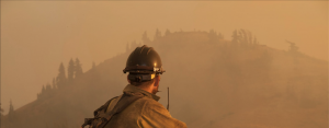 A fire fighter with a large pack, helmet & yellow jacket works on a fire.